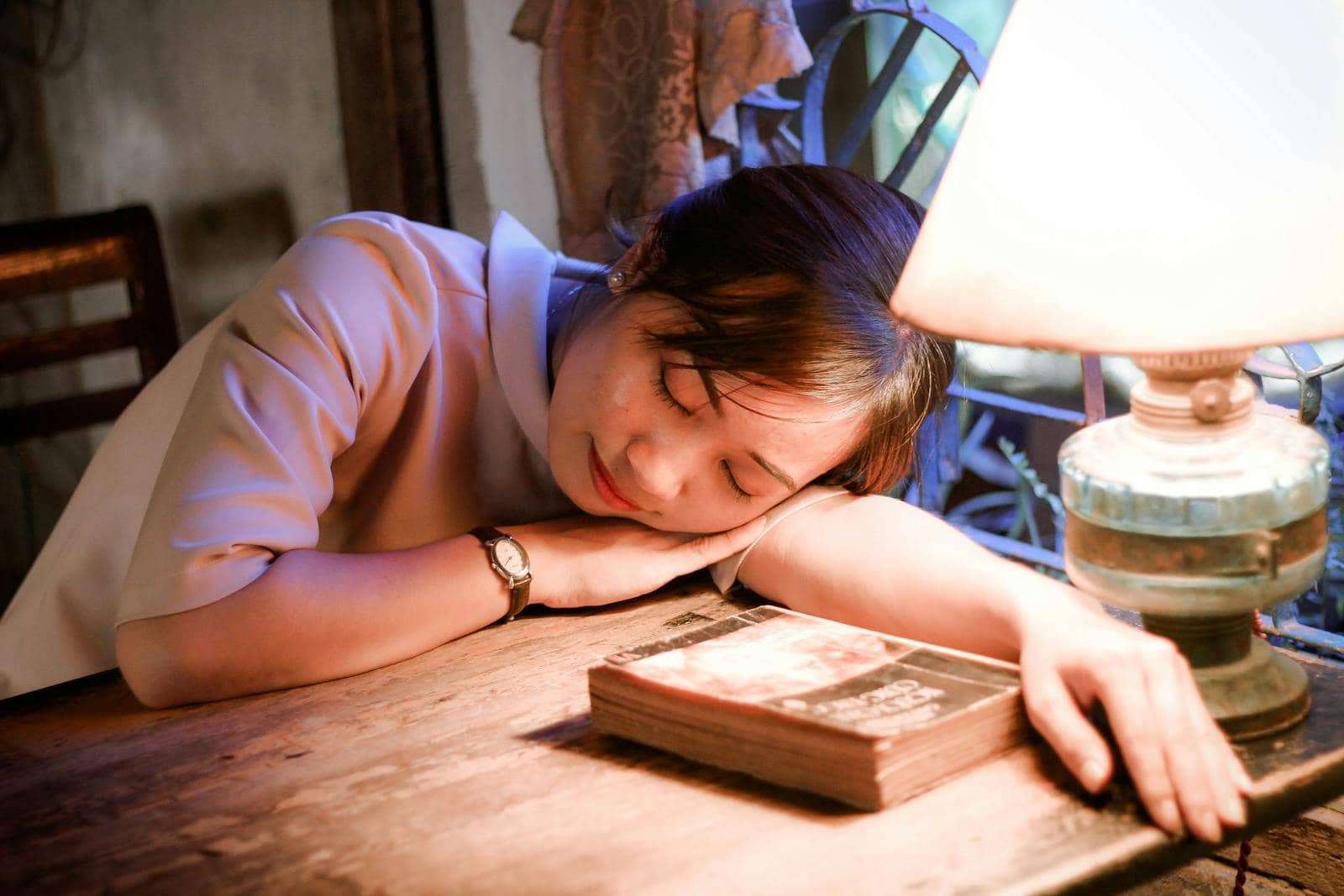 A young woman resting her head on a table under the warm glow of a lamp.